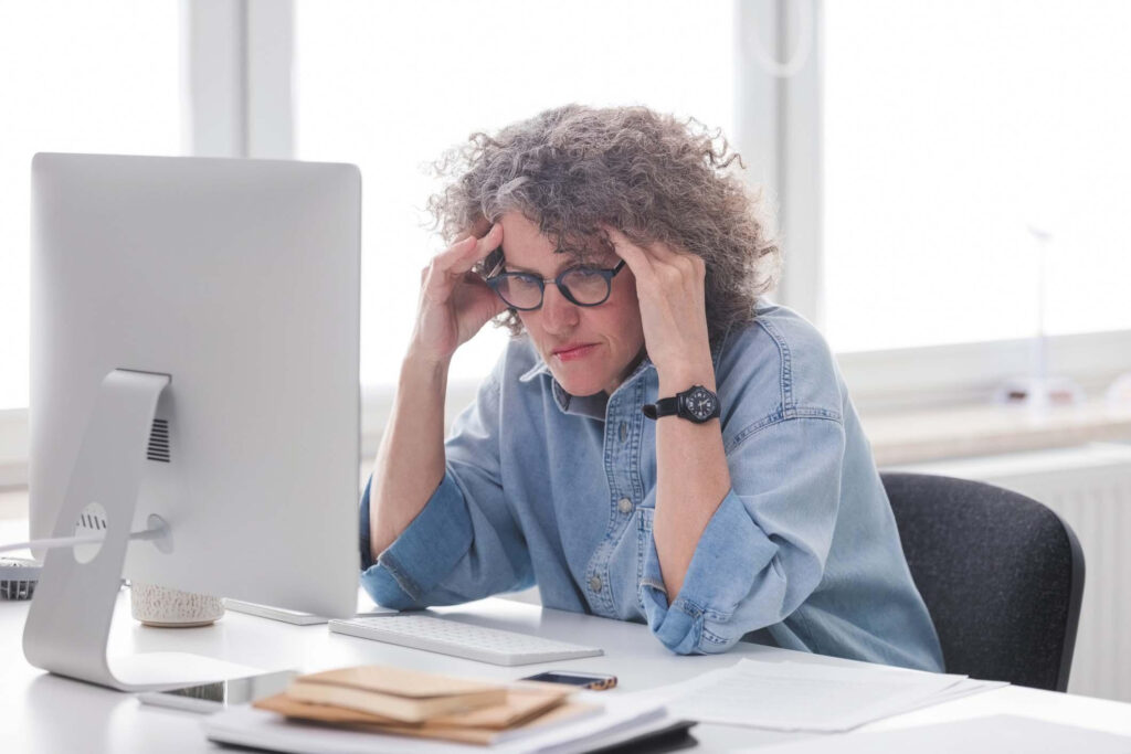 Una donna è seduta alla scrivania davanti a un computer, ha le mani ai lati della testa e sembra stressata.